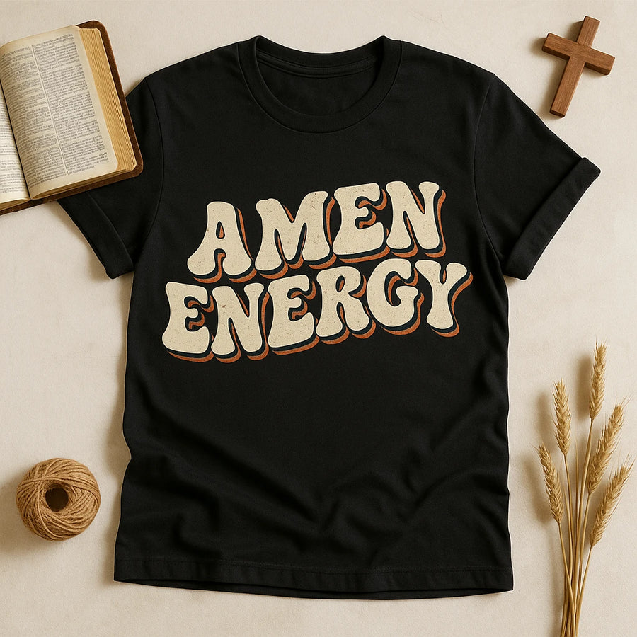 A black t-shirt with the words "AMEN ENERGY" written in bold, stylized font. The shirt is laid flat on a beige surface, surrounded by an open book, a wooden cross, a roll of twine, and some wheat stalks.
