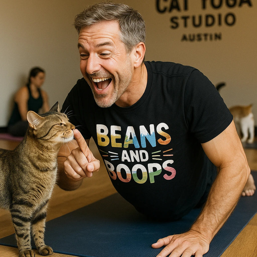 A man is lying on a yoga mat in a cat yoga studio, playfully interacting with a tabby cat. He is smiling and pointing his finger toward the cat. The man is wearing a black T-shirt with colorful letters that read "BEANS AND BOOPS." In the background, a woman is sitting on a yoga mat, and another cat is visible.