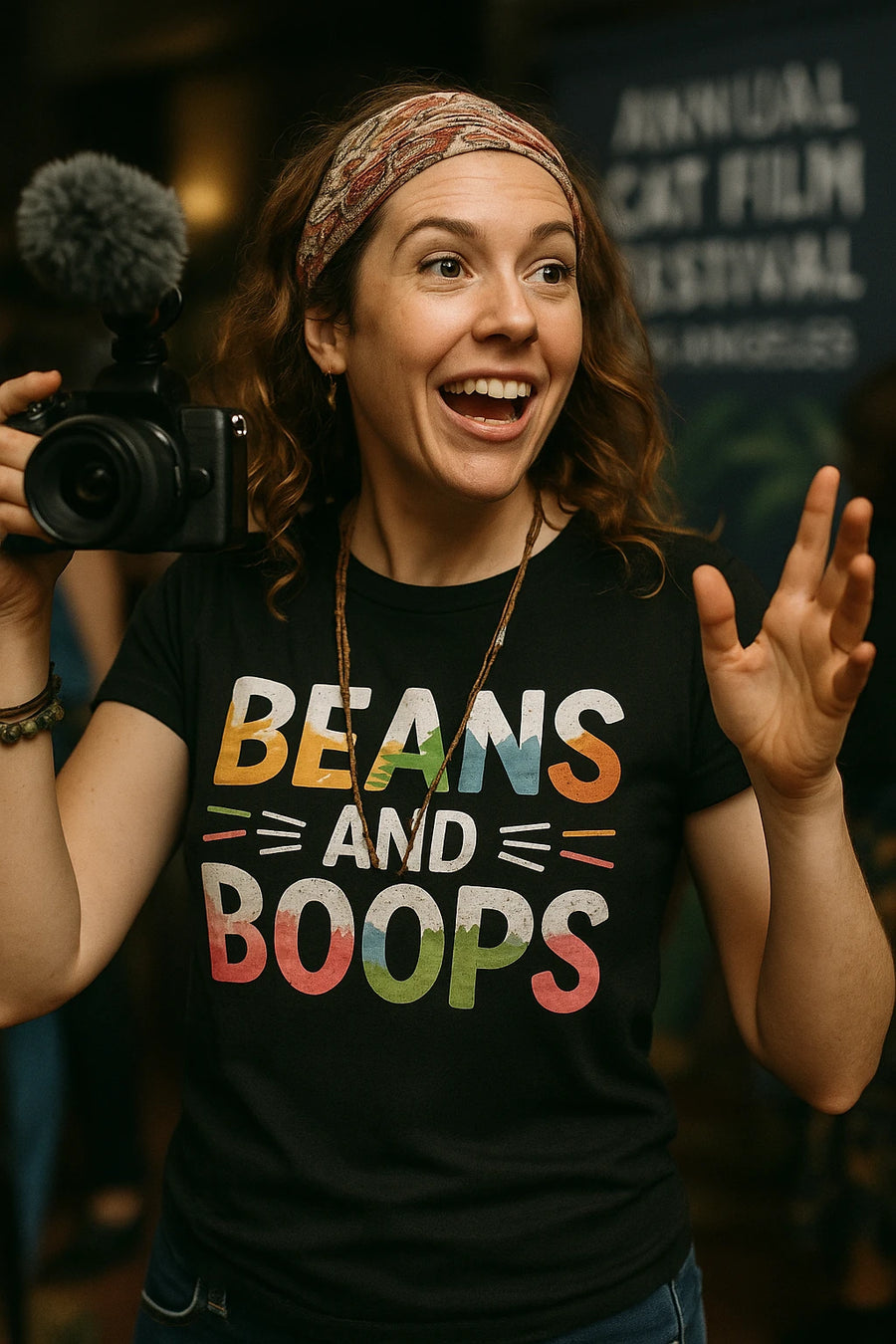 A person with curly hair, wearing a patterned headband and holding a camera with a fuzzy microphone. They are smiling widely, and wearing a black shirt with colorful text. In the background, there's a blurred sign that suggests a film festival event.