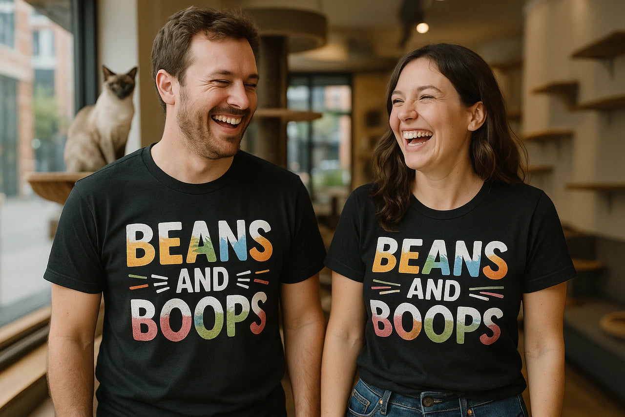 A man and a woman are standing indoors, both wearing black T-shirts with colorful text that reads "BEANS AND BOOPS." They are laughing and appear to be enjoying each other's company. A Siamese cat is perched on a ledge behind them. The setting seems to be a cozy, well-lit room with shelves and windows.
