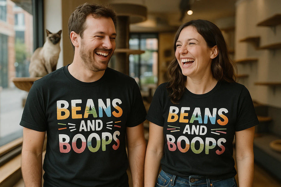 A man and a woman are standing indoors, both wearing black T-shirts with colorful text that reads "BEANS AND BOOPS." They are laughing and appear to be enjoying each other's company. A Siamese cat is perched on a ledge behind them. The setting seems to be a cozy, well-lit room with shelves and windows.