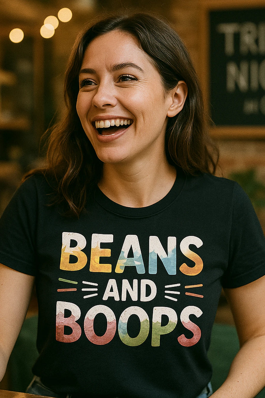 A person with long, dark hair is smiling broadly. They are wearing a black shirt with colorful text that reads "BEANS AND BOOPS." The background is softly blurred with warm lighting, suggesting a cozy indoor setting.