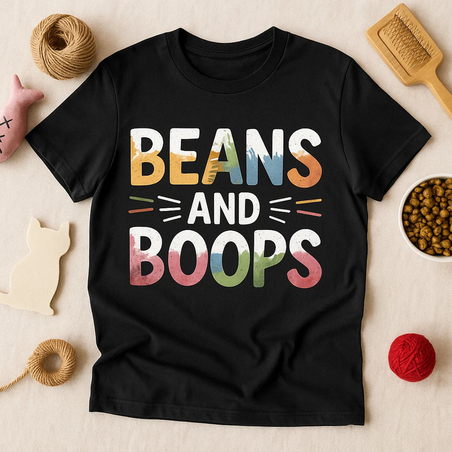 A black T-shirt with colorful, bold text that reads "BEANS AND BOOPS." The shirt is surrounded by various items, including a wooden brush, a bowl of kibble, yarn balls, and a cutout of a cat, against a beige background.