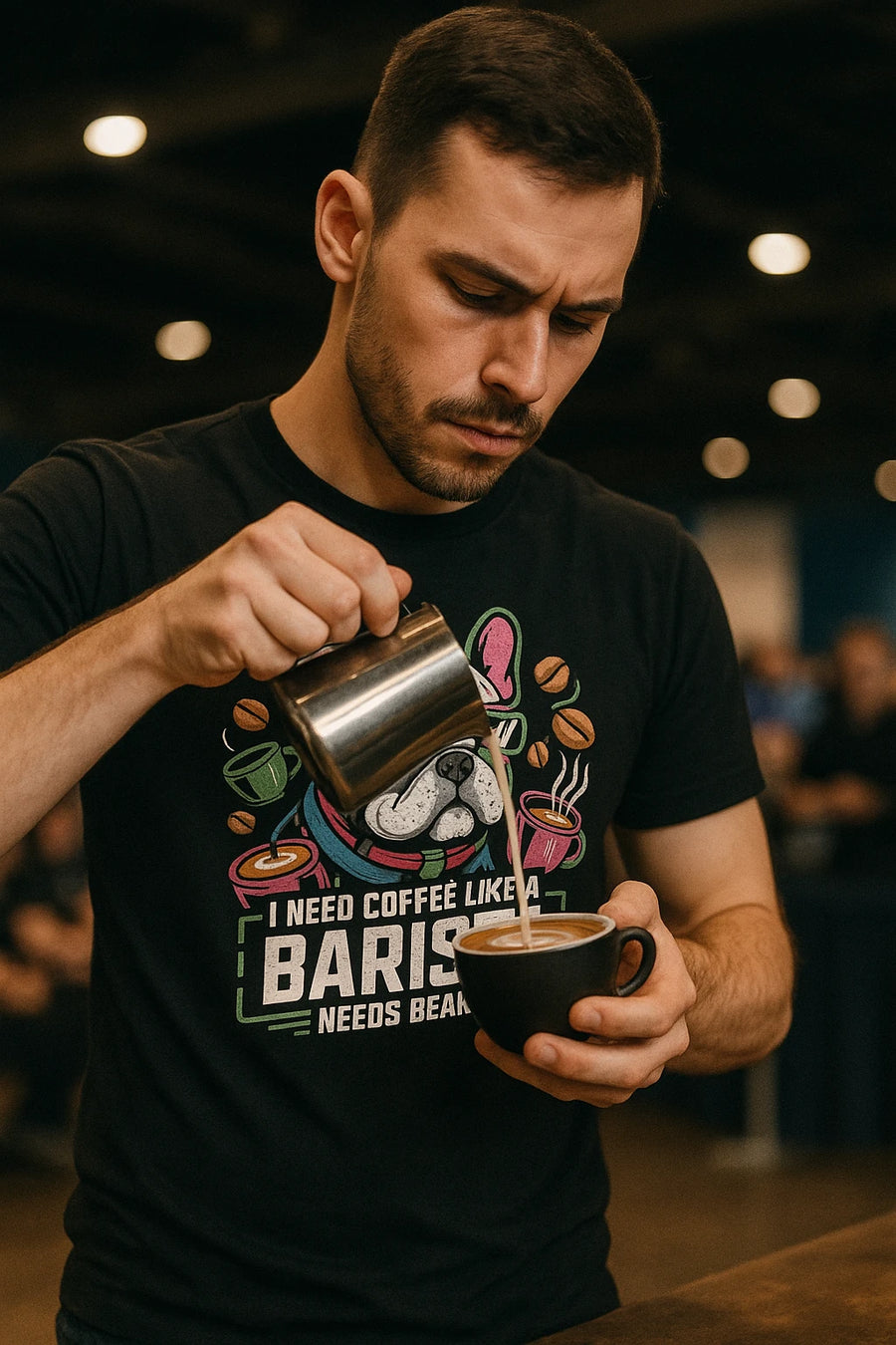 A person with short dark hair and a mustache is focused on pouring milk into a cup of coffee, creating latte art. They are wearing a black shirt with colorful graphics including a dog and coffee-related imagery. The background is a blurred indoor setting with warm lighting.
