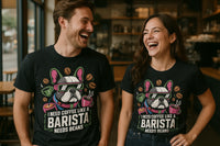 A man and a woman are standing in a coffee shop, laughing and wearing matching black t-shirts. The shirts have a colorful design of a dog with sunglasses surrounded by coffee beans and cups.
