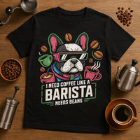 A black t-shirt featuring a graphic of a bulldog wearing sunglasses and surrounded by coffee cups and beans. The shirt is placed on a wooden surface with coffee-making utensils and coffee beans scattered around.