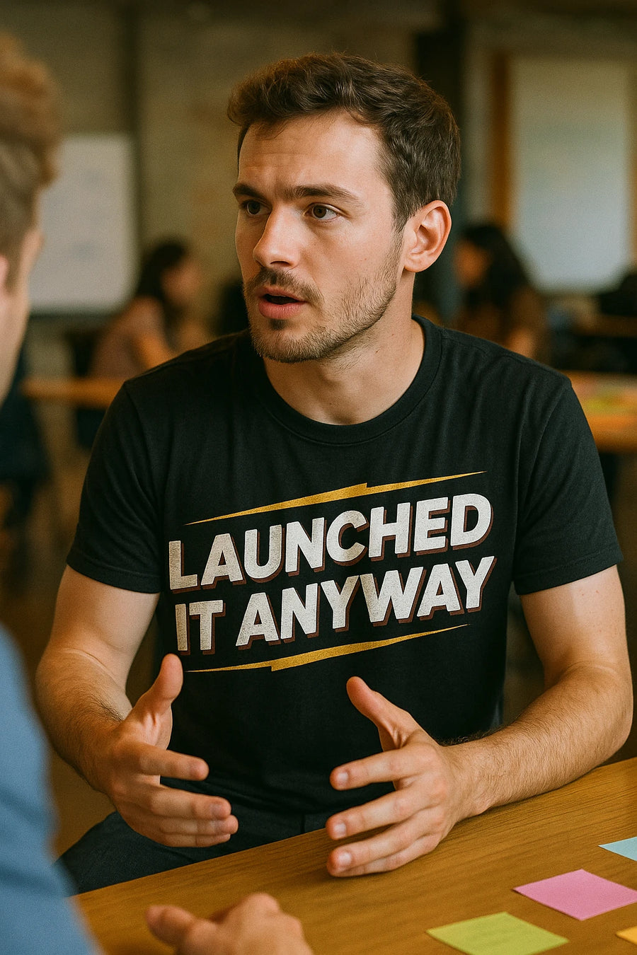 A person with short brown hair and facial hair is sitting at a table, engaged in conversation. They are wearing a black t-shirt with the text "LAUNCHED IT ANYWAY" printed in bold white letters with yellow accents. The background shows a blurred room with other people sitting at tables.