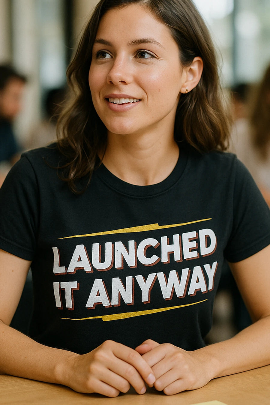 A person with shoulder-length brown hair wearing a black t-shirt with the text "LAUNCHED IT ANYWAY" sits at a table, smiling slightly. The background is softly blurred, suggesting an indoor setting.