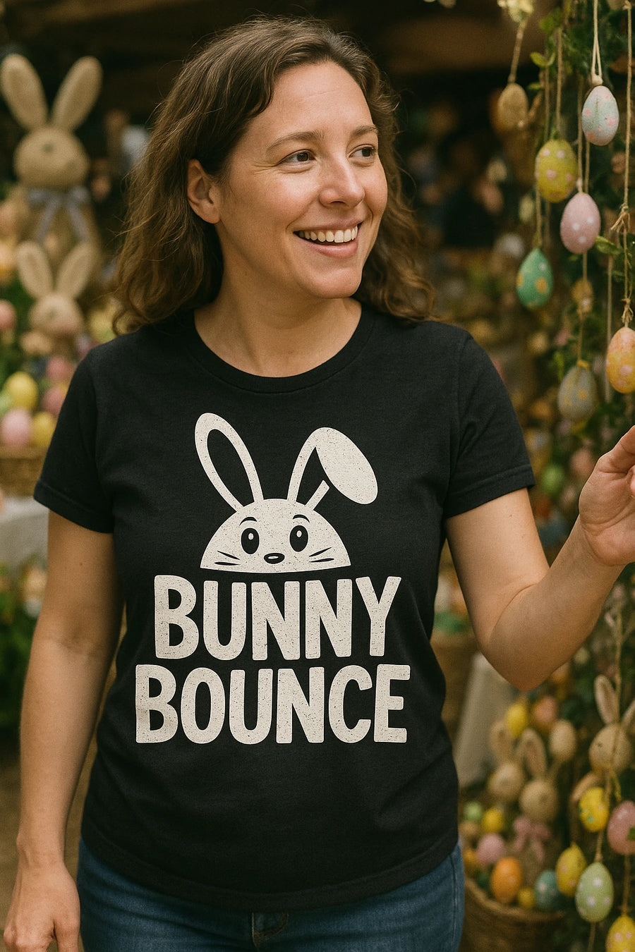 A person with shoulder-length brown hair is smiling while looking to the side. They are wearing a black shirt with a white graphic of a bunny head and the words "BUNNY BOUNCE" printed on it. The background features various decorative elements, including hanging eggs and bunny figures.