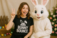 A person with long brown hair is smiling broadly and showing a peace sign with one hand while wearing a black t-shirt with a bunny graphic. Next to them is a person in a white bunny costume with large ears and a happy expression. The background includes Easter-themed decorations such as colorful eggs, flowers, and a wooden fence.