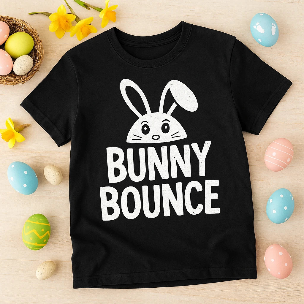 A black T-shirt with a cute cartoon bunny head graphic above the words "BUNNY BOUNCE" in white text. The shirt is laid out on a light wooden surface surrounded by colorful Easter eggs and yellow daffodil flowers.