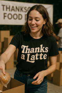 A person with shoulder-length brown hair and a black t-shirt is smiling while placing a wrapped item into a box. They are participating in a food drive, as indicated by a banner in the background that reads 