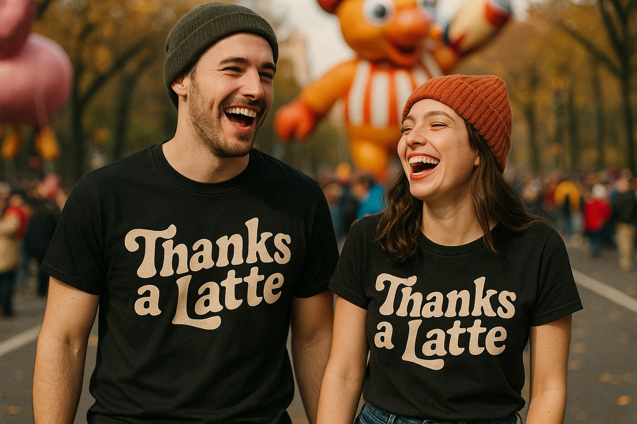 Two people, a man and a woman, are laughing together outdoors wearing matching black t-shirts with the text "Thanks a Latte." The man is wearing a green beanie and the woman is wearing a red beanie. There is a large balloon figure and a crowd in the background, suggesting a festive event or parade.
