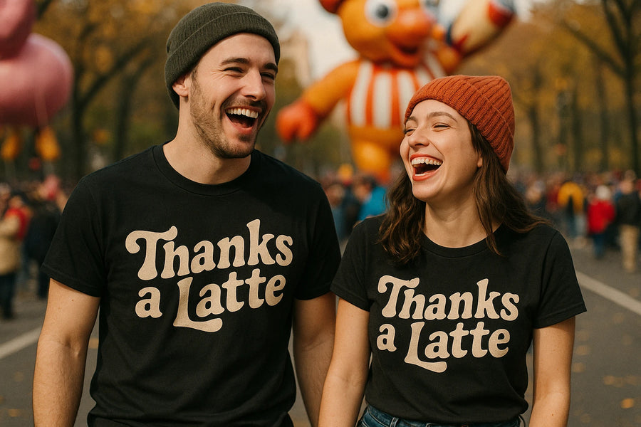 Two people, a man and a woman, are laughing together outdoors wearing matching black t-shirts with the text "Thanks a Latte." The man is wearing a green beanie and the woman is wearing a red beanie. There is a large balloon figure and a crowd in the background, suggesting a festive event or parade.