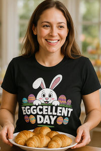 A person with long brown hair is smiling and holding a plate of croissants. They are wearing a black t-shirt featuring a cute cartoon bunny peeking over decorated Easter eggs, with the text 