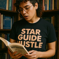 A person with curly hair and glasses is standing in front of a bookshelf, holding an open book titled 