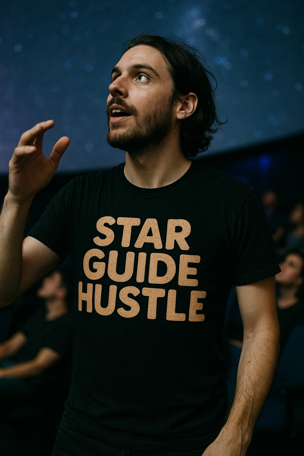 A person with a beard and long hair is gazing upwards, holding one hand raised. They are wearing a black T-shirt with the phrase "STAR GUIDE HUSTLE" printed in bold letters. The background suggests a setting similar to a planetarium, with people blurred in the distance.