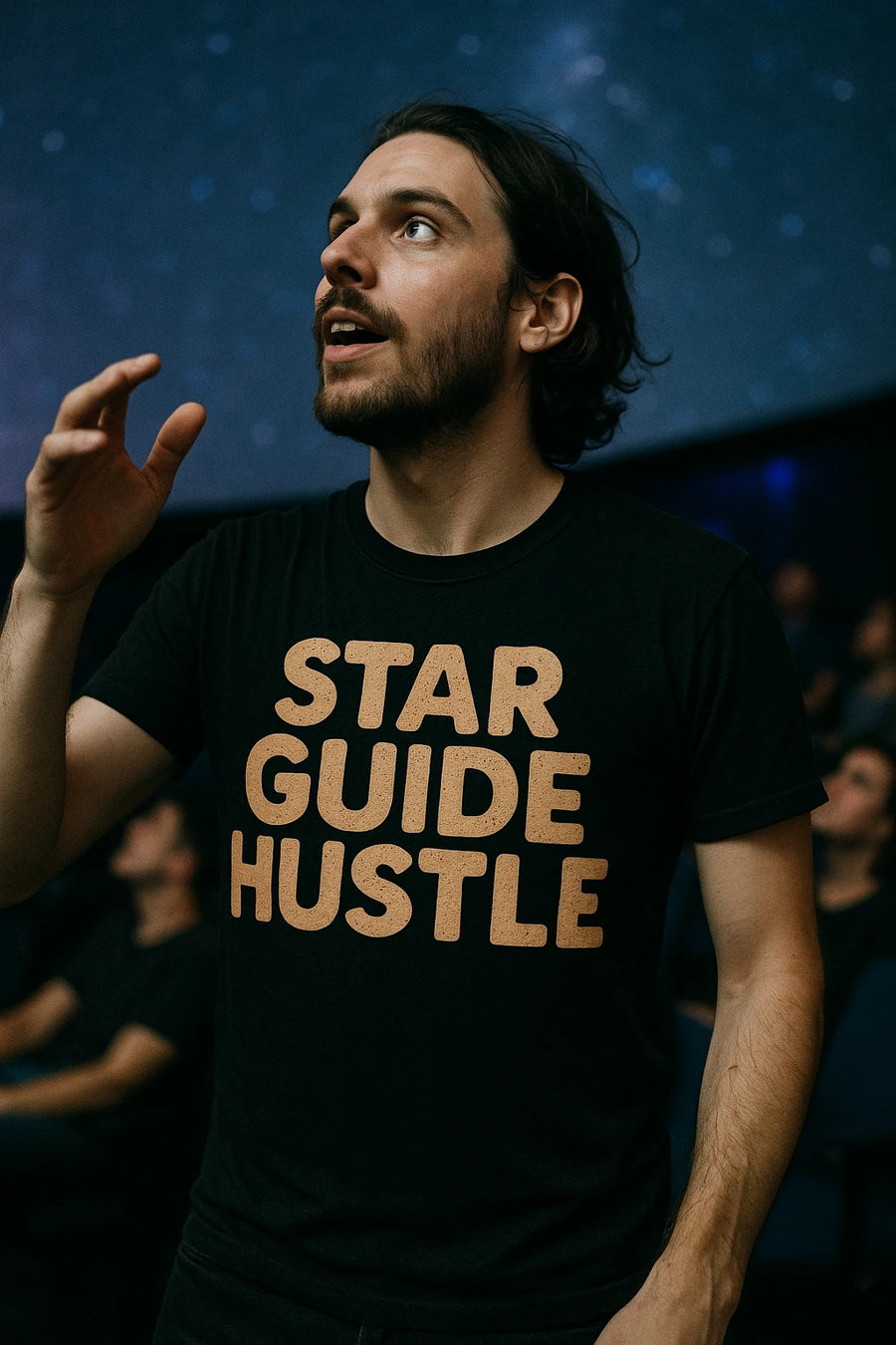 A person with a beard and long hair is gazing upwards, holding one hand raised. They are wearing a black T-shirt with the phrase "STAR GUIDE HUSTLE" printed in bold letters. The background suggests a setting similar to a planetarium, with people blurred in the distance.