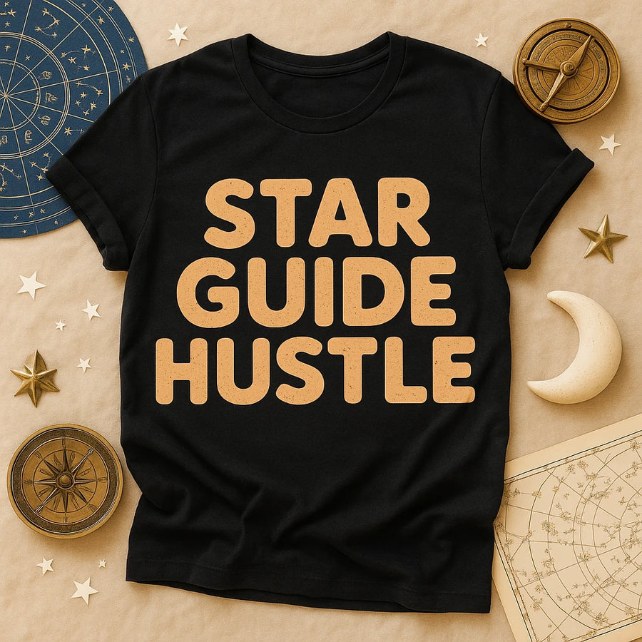 A black t-shirt with the words "STAR GUIDE HUSTLE" in large, bold, beige letters. The shirt is laid out flat on a beige surface, surrounded by celestial-themed items including a map with constellations, a compass, a crescent moon, and decorative stars.