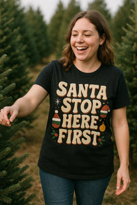 A person with shoulder-length brown hair is smiling and standing among evergreen trees. They are wearing a black T-shirt with festive decorations and playful text. The background shows a Christmas tree farm setting.