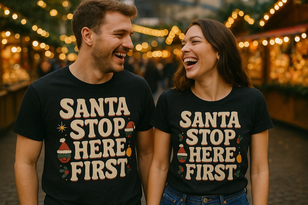 A man and woman are laughing together in a festive setting with warm, glowing lights in the background. Both are wearing matching black T-shirts that say "Santa Stop Here First." The scene suggests a cheerful holiday atmosphere.
