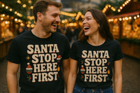 A man and woman are laughing together in a festive setting with warm, glowing lights in the background. Both are wearing matching black T-shirts that say 