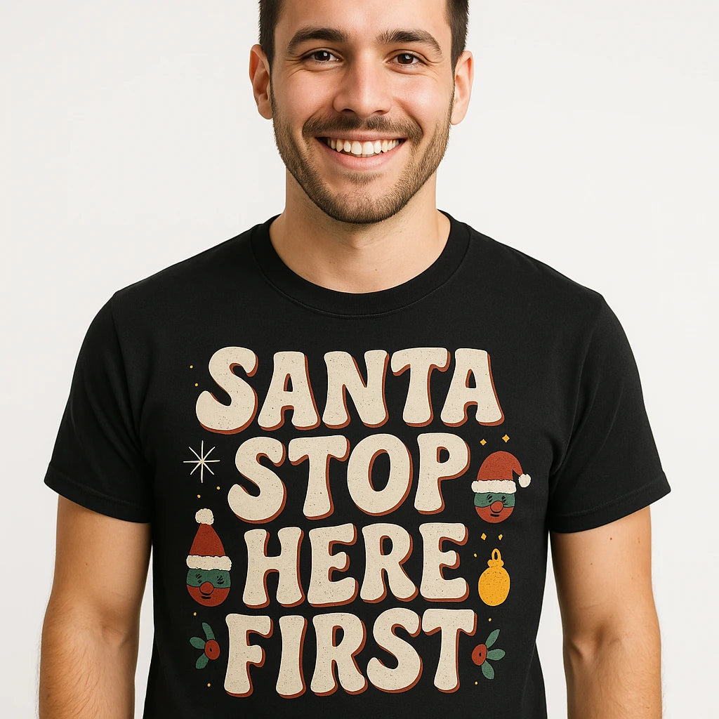 A person is smiling while wearing a black shirt with the text "SANTA STOP HERE FIRST" in large, festive letters. The design includes small holiday-themed graphics, such as a Santa hat and ornaments.