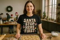 A person is smiling at the camera, standing in a kitchen with a tray of cookie dough shaped into stars in front of them. They are wearing a black t-shirt with the text “SANTA STOP HERE FIRST” and holiday decorations printed on it. In the background, several people wearing Santa hats are engaged in baking activities. A wreath is on the wall, and there are festive decorations on the counter.