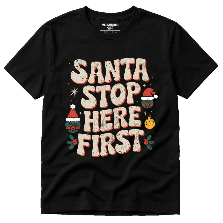 A black T-shirt with a festive design that includes the phrase "Santa Stop Here First" in large, bold letters. The text is decorated with holiday-themed graphics, such as Santa hats, ornaments, and holly.