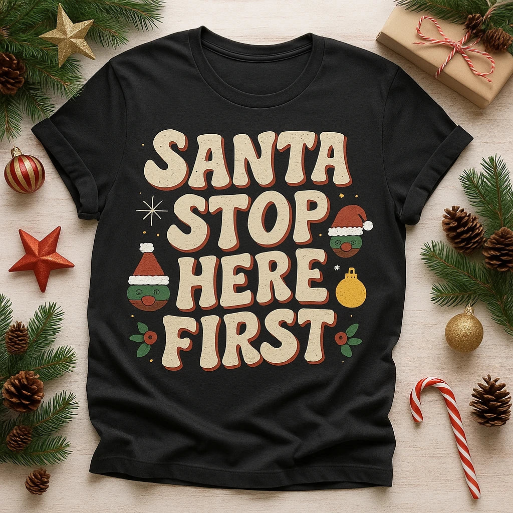 A black t-shirt with festive lettering that says "SANTA STOP HERE FIRST" in cream and red colors. The design includes small illustrations of a Christmas hat and ornaments. The t-shirt is surrounded by Christmas decorations, including pine branches, pine cones, a candy cane, and wrapped gift boxes.