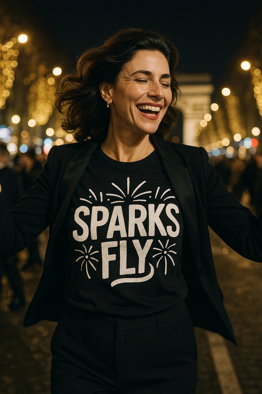A person with shoulder-length dark hair is joyfully smiling with their eyes closed. They are wearing a black jacket over a shirt with the words "SPARKS FLY" written on it, surrounded by firework designs. The background features a blurred night scene with bright lights, possibly on a city street.