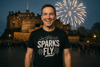 A smiling person stands in front of a historical castle at night, with fireworks lighting up the sky in the background. The person is wearing a black t-shirt with the text 