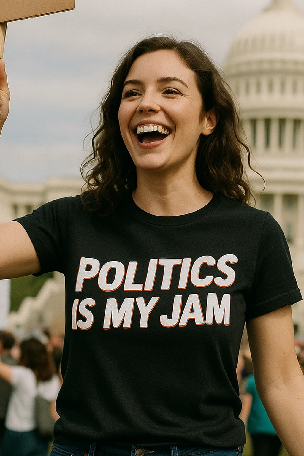 A person with long, wavy hair is smiling broadly while wearing a black shirt with the text "POLITICS IS MY JAM." They are standing outdoors, with a recognizable domed building in the background, likely indicating a political event or protest. The atmosphere appears lively.