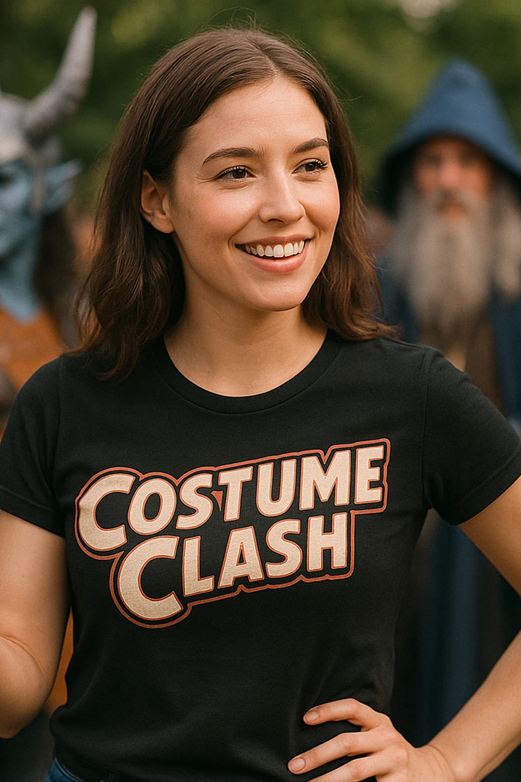 A person with long brown hair is smiling while wearing a black T-shirt with "COSTUME CLASH" printed on it. In the background, a person in a blue hood and a costumed figure with horns can be seen, both out of focus.