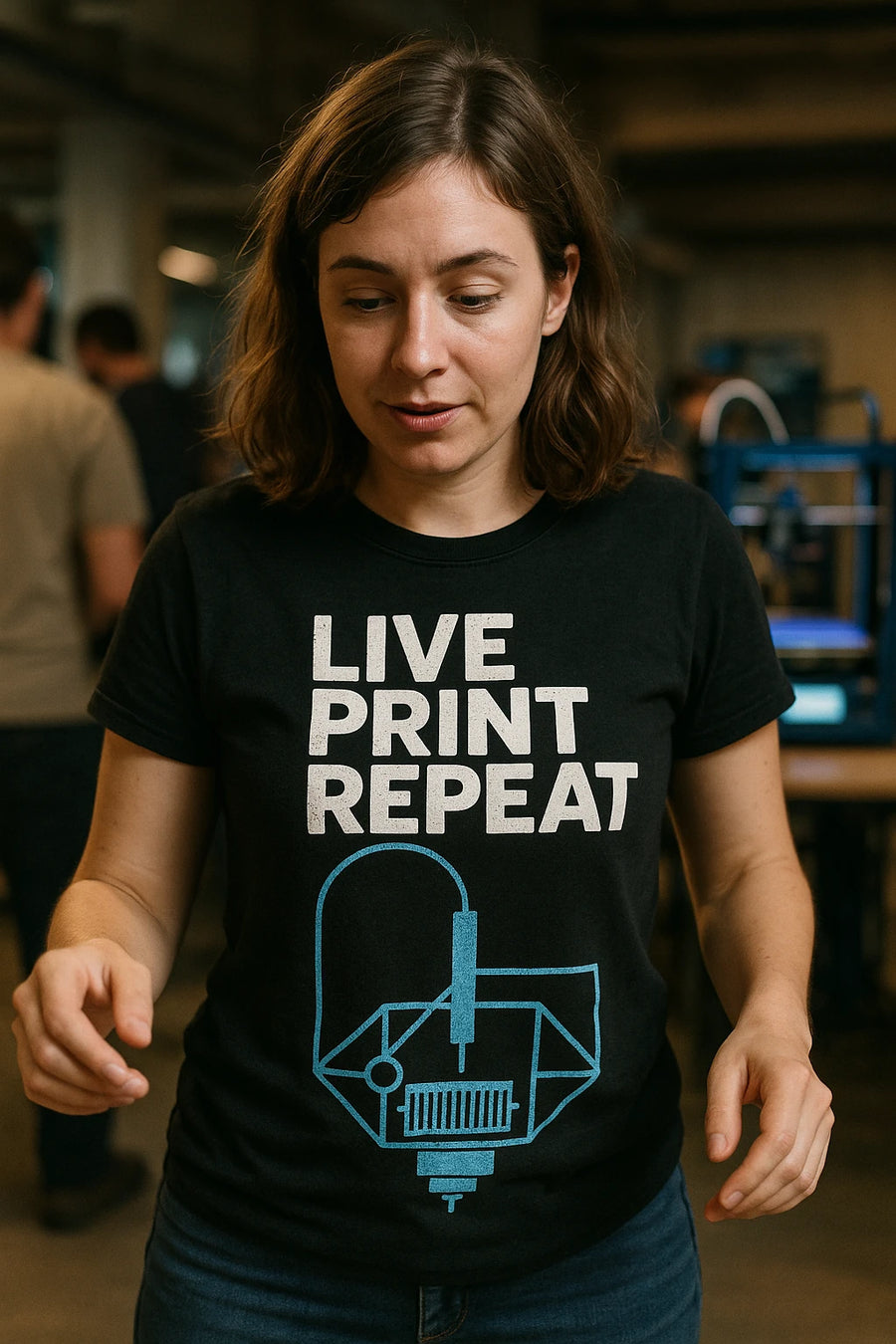 A person with shoulder-length brown hair is wearing a black t-shirt with the text "LIVE PRINT REPEAT" and a design of a 3D printer. They are standing in a room with other people and 3D printers in the background.