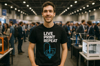 A person wearing a black T-shirt with white and blue text stands in a busy, indoor event or conference setting. There are multiple people in the background, along with tables and equipment, creating a bustling atmosphere.