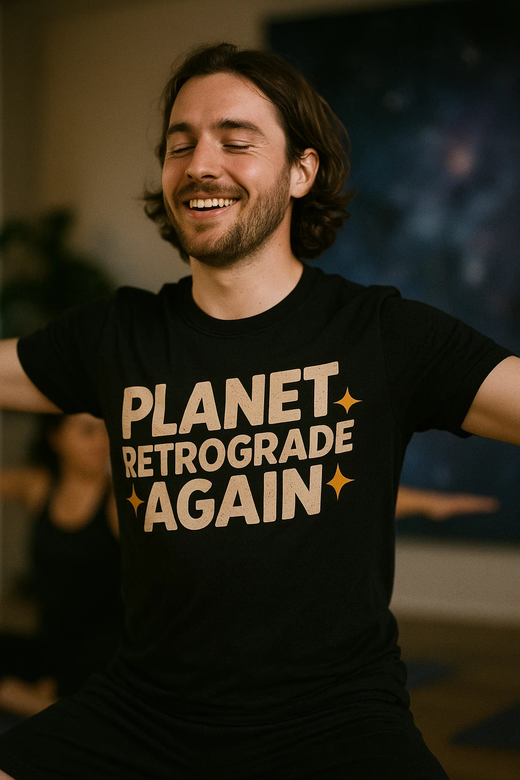 A person with long hair and a beard is sitting with arms outstretched, smiling with eyes closed, wearing a black T-shirt with the text "PLANET RETROGRADE AGAIN" in bold letters.