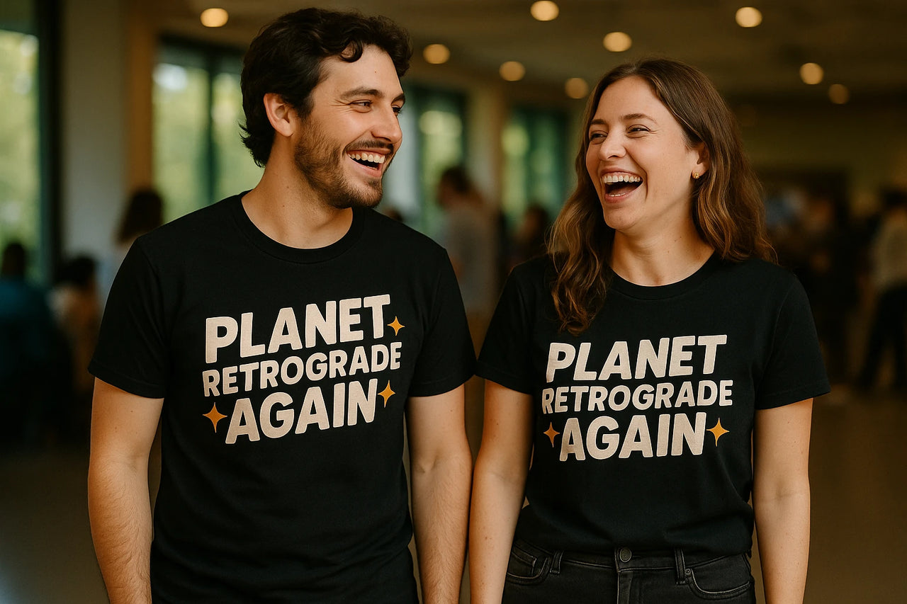 A man and a woman are standing side by side, both wearing black t-shirts with the text "PLANET RETROGRADE AGAIN" printed on them. They are laughing and looking at each other, set against a blurred indoor background with soft lighting.