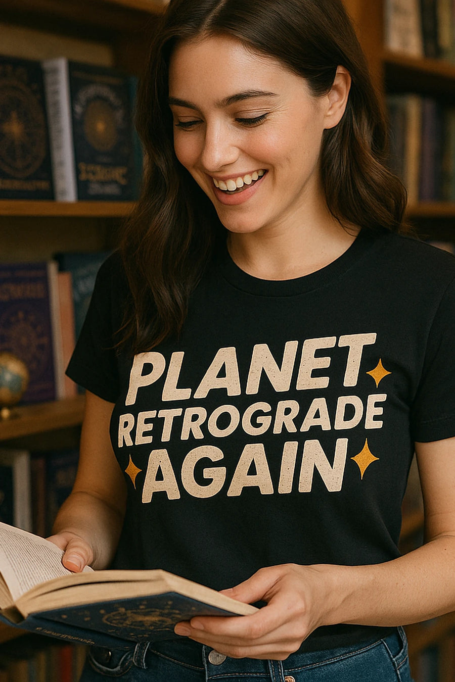 A person with long brown hair is smiling while holding an open book. They are wearing a black t-shirt with the text "PLANET RETROGRADE AGAIN." Shelves filled with books are visible in the background.