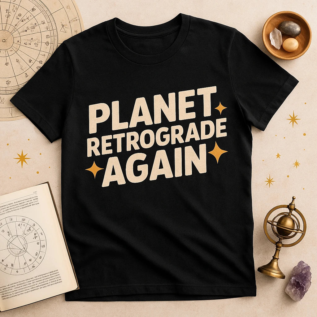 A black t-shirt with bold white text saying "PLANET RETROGRADE AGAIN" with decorative stars. The shirt is laid out on a surface surrounded by astrological items such as zodiac charts, a small globe, and crystals.