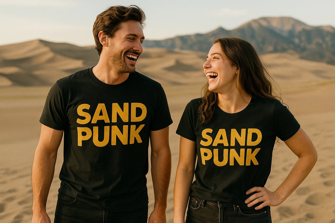A man and a woman are standing side by side in a desert landscape, laughing with each other. They are both wearing black t-shirts with the words "SAND PUNK" in large yellow letters. The background features sand dunes and distant mountains.