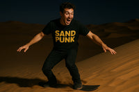 A person with curly hair is enthusiastically sandboarding down a dune at night. They are wearing a black t-shirt with 