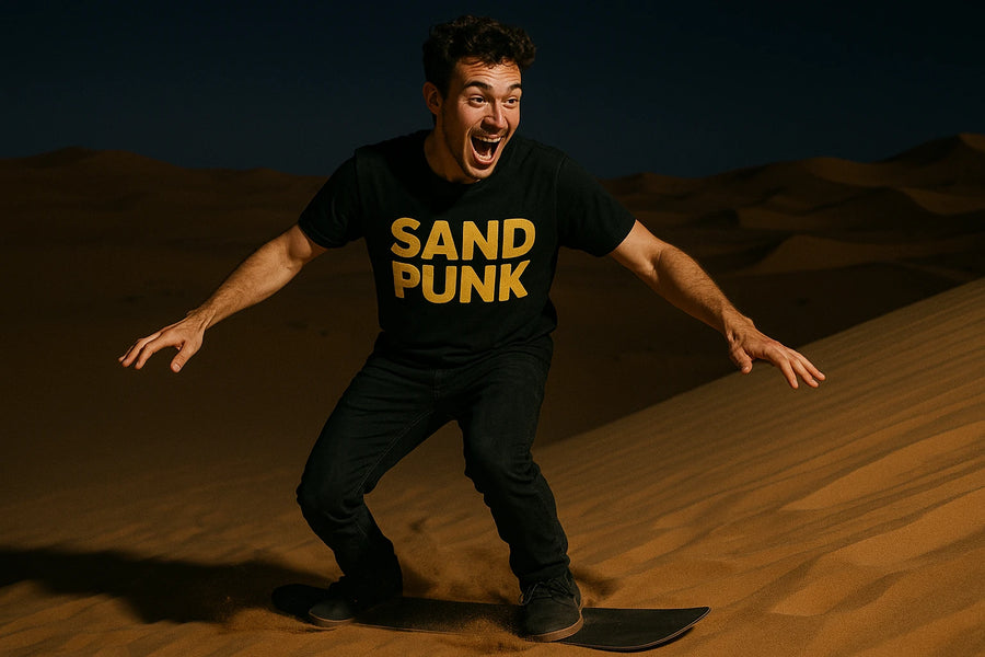 A person with curly hair is enthusiastically sandboarding down a dune at night. They are wearing a black t-shirt with "SAND PUNK" written in bold yellow letters, black pants, and dark shoes. The background shows the silhouette of sand dunes under a dark sky.