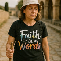 A person with closed eyes stands on a cobblestone path, wearing a black T-shirt with colorful text. They have shoulder-length hair and are wearing a beige sun hat, carrying a wooden walking stick and a black backpack. Stone arches and greenery are visible in the background.