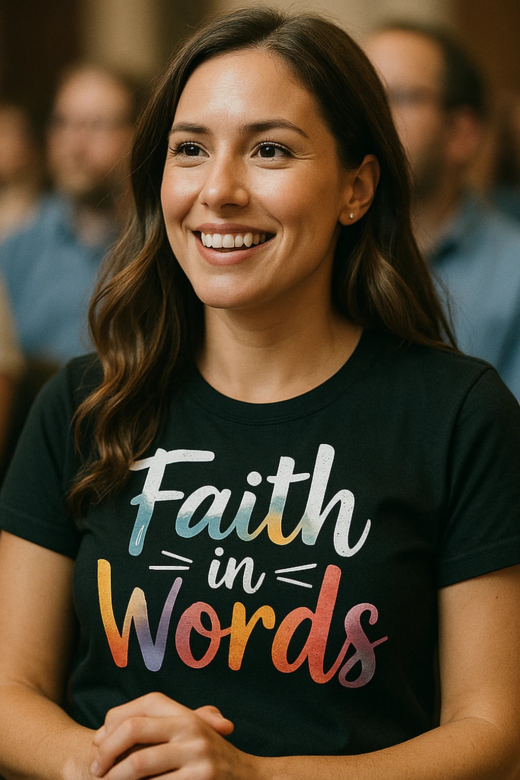 A person with long brown hair is smiling and wearing a black t-shirt with colorful text that reads "Faith in Words." They are surrounded by a blurred background with other people.
