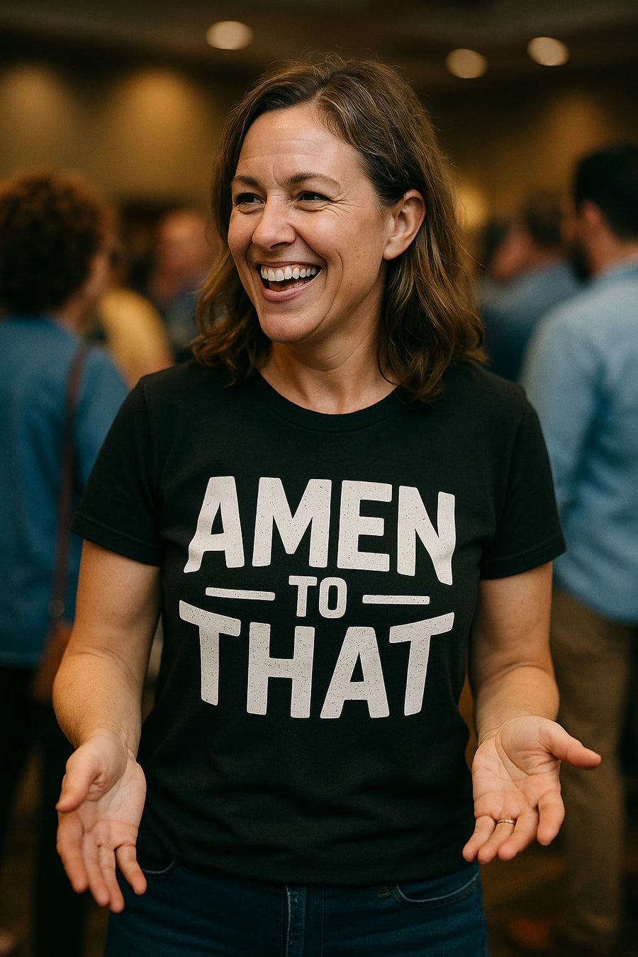 A person with shoulder-length brown hair is smiling broadly in a crowded room. They are wearing a black T-shirt with the text "AMEN TO THAT" printed in bold white letters. The background shows several other people out of focus.