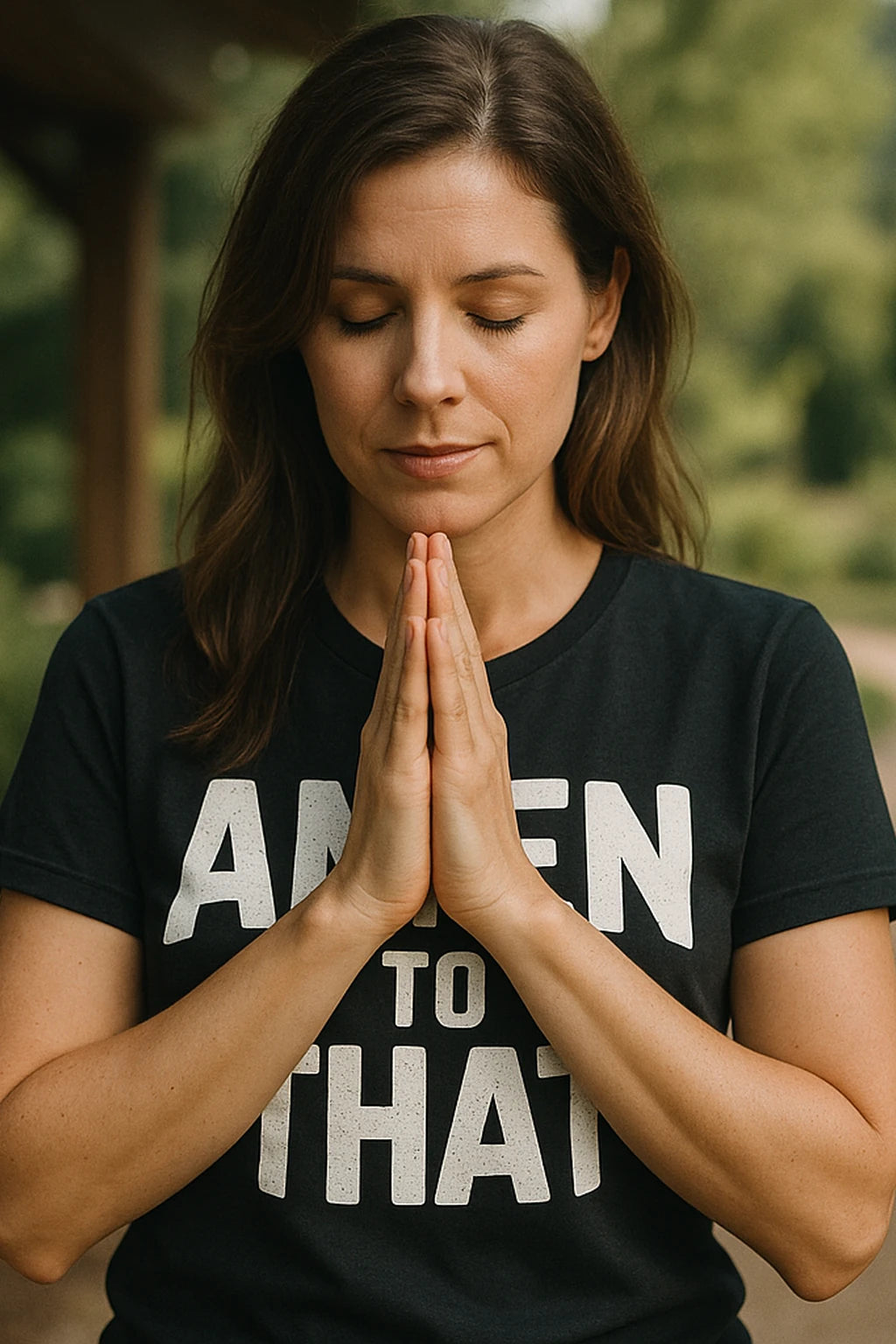 A person with long brown hair is standing outdoors with their eyes closed and hands pressed together in a prayer-like position. They are wearing a black T-shirt with white text.