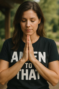 A person with long brown hair is standing outdoors with their eyes closed and hands pressed together in a prayer-like position. They are wearing a black T-shirt with white text.