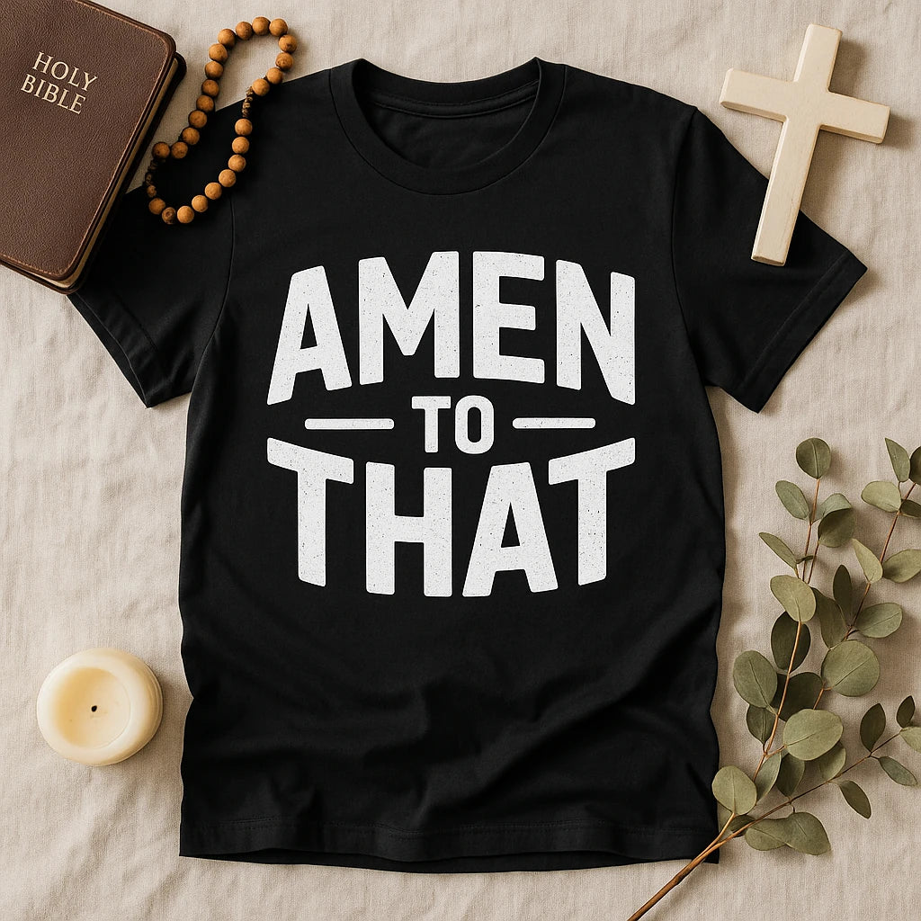 A black t-shirt with the text "AMEN TO THAT" in bold white letters. The shirt is laid on a beige surface surrounded by a Bible, a rosary, a cross, a candle, and some greenery, creating a spiritual-themed arrangement.