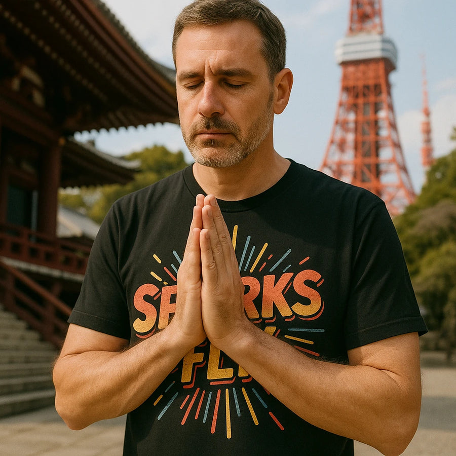 A person with short hair and a beard is standing outdoors with their eyes closed, hands pressed together in a meditative pose. They are wearing a black T-shirt with colorful text that reads "SPARKS FLY." In the background, there is a traditional wooden structure and a tall tower.